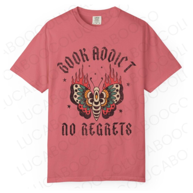 May include: A dusty rose t-shirt with the words "BOOK ADDICT" above a butterfly design with flames and the words "NO REGRETS" below. The butterfly has red, green, and yellow details.