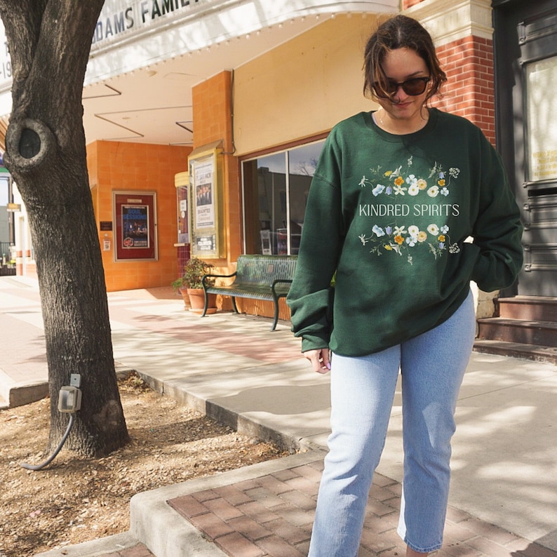 May include: A dark green sweatshirt with a floral design and the text "KINDRED SPIRITS" in white. The person wearing the sweatshirt is standing on a sidewalk in front of a building.