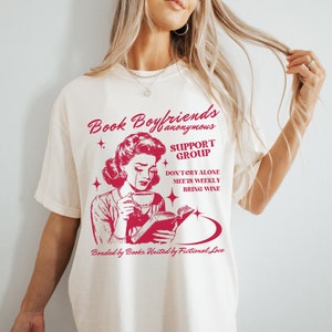 May include: White t-shirt with a red graphic of a woman reading a book and crying. The text on the shirt reads "Book Boyfriends anonymous SUPPORT GROUP DON'T CRY ALONE MEETS WEEKLY BRING WINE Bonded by Books. United by Fictional Love."