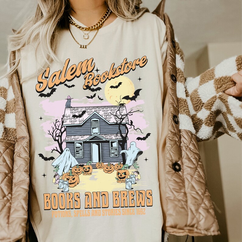 Op de afbeelding: Een beige T-shirt met een grafische print van een spookhuis, vleermuizen en een volle maan. De tekst "Salem Bookstore" staat in een retro-lettertype boven het huis. Onder het huis staat de tekst "Books and Brews" met de slogan "Potions, Spells and Stories Since 1992".