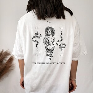 May include: White t-shirt with a black and white graphic print of a woman with snakes and the text "STRENGTH BEAUTY POWER".