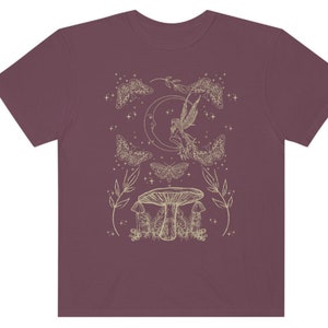 Pu&ograve; includere: Una t-shirt color granata con un contorno bianco di una fata, una luna crescente, falene e funghi. Il design &egrave; in stile fantasioso.