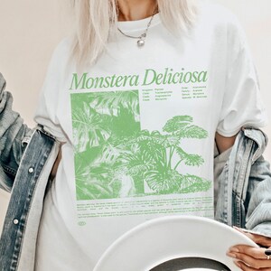 May include: White t-shirt with a green illustration of a Monstera Deliciosa plant. The shirt has the scientific classification of the plant printed on it, including the kingdom, class, order, family, genus, and species. The text "Monstera Deliciosa" is printed in large, bold letters at the top of the shirt.