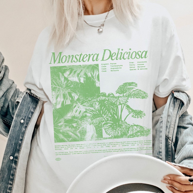 May include: White t-shirt with a green illustration of a Monstera Deliciosa plant. The shirt has the scientific classification of the plant printed on it, including the kingdom, class, order, family, genus, and species. The text "Monstera Deliciosa" is printed in large, bold letters at the top of the shirt.