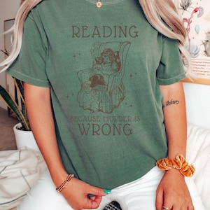 Puede incluir: Una camiseta verde con una ilustraci&oacute;n vintage de una persona leyendo en una silla. El texto en la camiseta dice "Reading because murder is wrong."