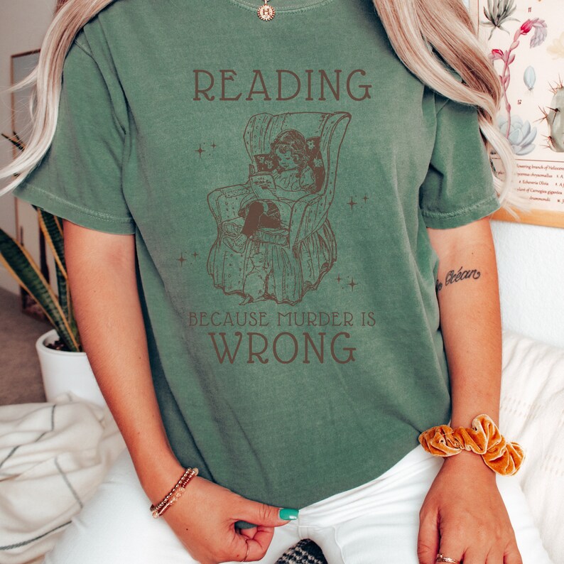 Puede incluir: Una camiseta verde con una ilustraci&oacute;n vintage de una persona leyendo en una silla. El texto en la camiseta dice "Reading because murder is wrong."