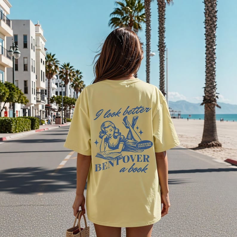 K&ouml;nnte beinhalten: Hellgelbes T-Shirt mit blauem Text "I look better bent over a book". Die Grafik zeigt eine Vintage-Illustration einer lesenden Frau. Das Shirt ist oversized und hat kurze &Auml;rmel.
