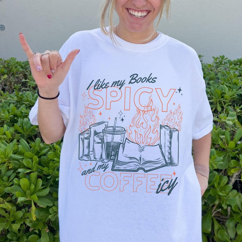 May include: White t-shirt with the text "I like my Books SPICY and my COFFEE icy." The graphic includes illustrations of books, a coffee drink, and flames. The design is in black and orange.