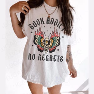 May include: White t-shirt with a graphic design featuring a butterfly with flames and the text "BOOK ADDICT NO REGRETS". The butterfly has teal, pink, yellow, and black details. The shirt is a casual style.
