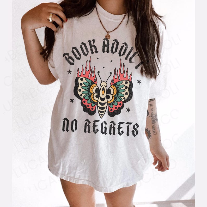 May include: White t-shirt with a graphic design featuring a butterfly with flames and the text "BOOK ADDICT NO REGRETS". The butterfly has teal, pink, yellow, and black details. The shirt is a casual style.