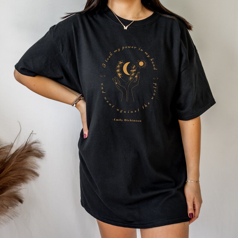 May include: Black oversized t-shirt with a gold graphic of hands holding a crescent moon and sun with a quote from Emily Dickinson: "I took my power in my hand and went against the world."