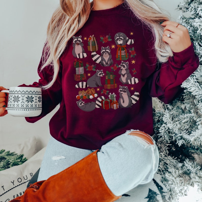 K&ouml;nnte beinhalten: Ein weinroter Pullover mit einem festlichen Design, das Waschb&auml;ren zeigt, die Geschenke halten und von Weihnachtsdekorationen umgeben sind.