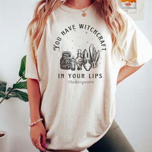 Op de afbeelding: Een wit T-shirt met een zwart grafisch ontwerp met de tekst "You Have Witchcraft In Your Lips -Shakespeare". De grafiek toont een cirkel met sterren, kristallen en flesjes met toverdrank.