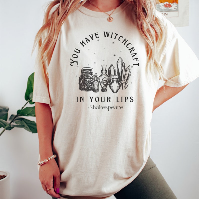 Op de afbeelding: Een wit T-shirt met een zwart grafisch ontwerp met de tekst "You Have Witchcraft In Your Lips -Shakespeare". De grafiek toont een cirkel met sterren, kristallen en flesjes met toverdrank.