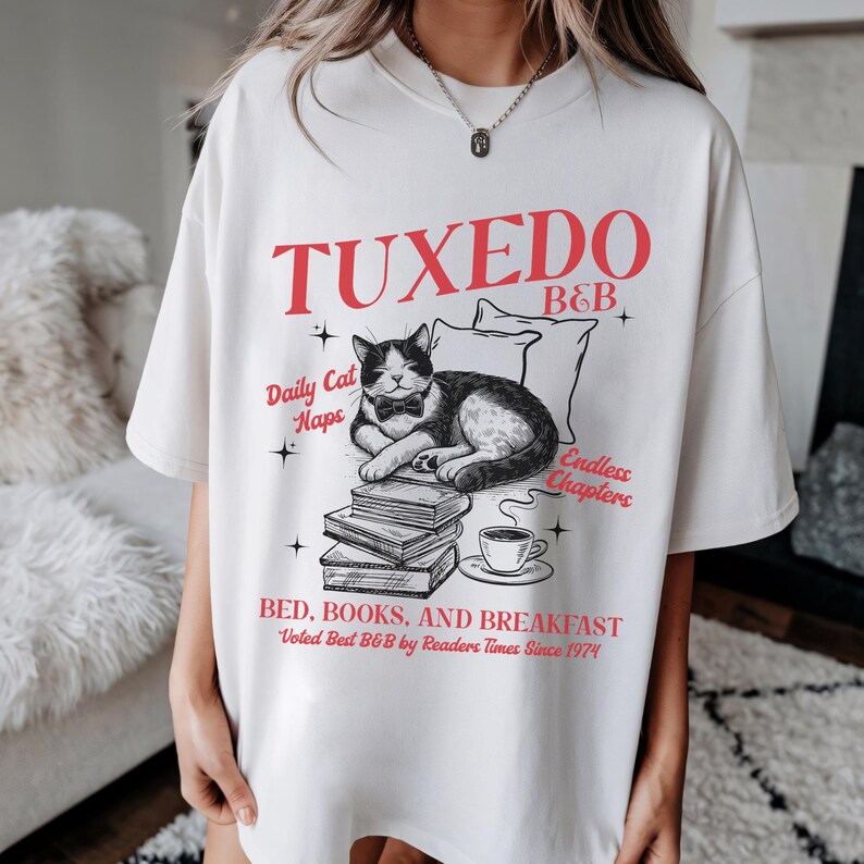 Op de afbeelding: Wit T-shirt met een zwart-witte illustratie van een kat met een strikje die slaapt op een stapel boeken. De tekst "TUXEDO B&B" staat in rood boven de kat en de tekst "Daily Cat Naps" en "Endless Chapters" staat in zwart onder de kat. De tekst "BED, BOOKS, AND BREAKFAST" staat in rood onder de boeken en de tekst "Voted Best B&B by Readers Times Since 1974" staat in zwart daaronder.