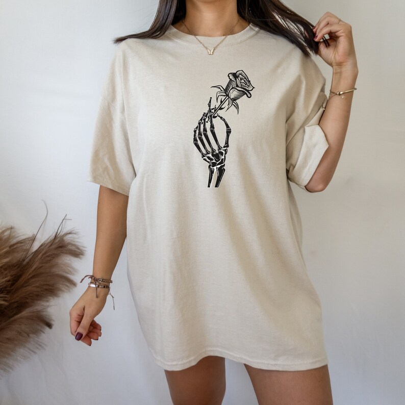 K&ouml;nnte beinhalten: Ein beigefarbenes Oversize-T-Shirt mit einem schwarzen Skelett-Hand-Motiv, das eine Rose h&auml;lt.