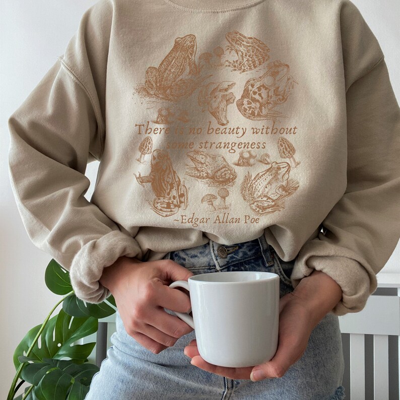 May include: A beige sweatshirt with a brown graphic print of frogs and mushrooms. The text "There is no beauty without some strangeness - Edgar Allan Poe" is printed on the sweatshirt.