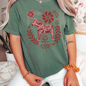May include: A green t-shirt featuring a red and white Scandinavian Dala horse design. The horse is surrounded by red flowers and leaves.