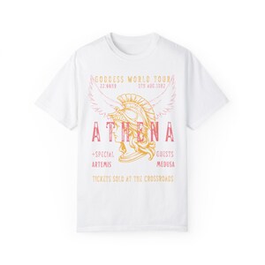 Puede incluir: Camiseta blanca con un dise&ntilde;o gr&aacute;fico de una mujer con un casco con alas. El texto dice "GODDESS WORLD TOUR", "ATHENA", "+SPECIAL ARTEMIS", "GUESTS MEDUSA", y "TICKETS SOLD AT THE CROSSROADS".