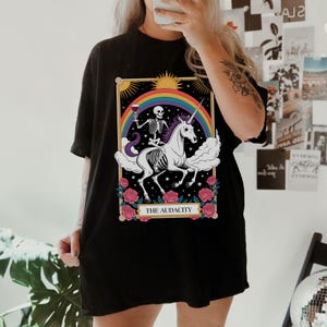 May include: Black t-shirt featuring a tarot card design. The graphic depicts a skeleton riding a unicorn under a rainbow, holding a wine glass. The card is framed with roses and the text "THE AUDACITY".