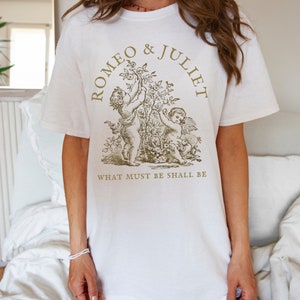 May include: White t-shirt with a gold graphic of two cherubs holding a rose bush. The text "ROMEO & JULIET" is above the cherubs and "WHAT MUST BE SHALL BE" is below.