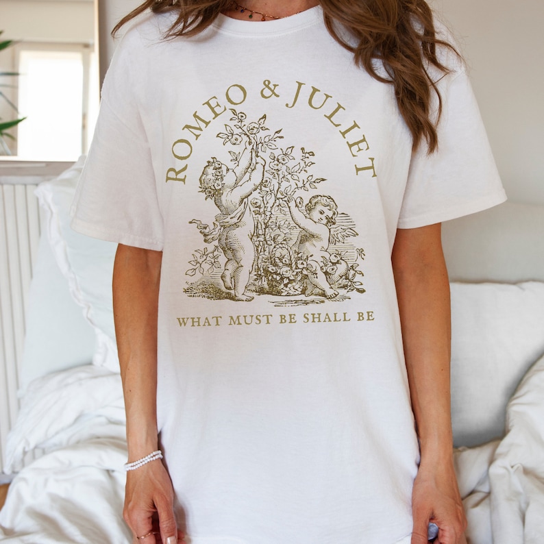 May include: White t-shirt with a gold graphic of two cherubs holding a rose bush. The text "ROMEO & JULIET" is above the cherubs and "WHAT MUST BE SHALL BE" is below.
