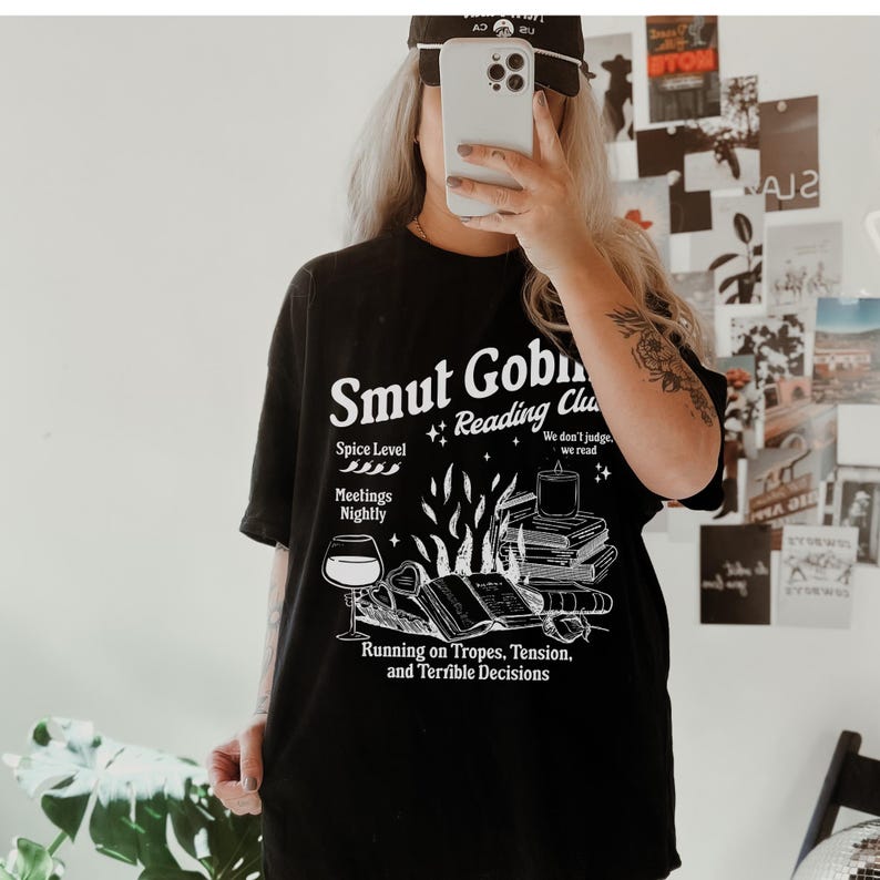 May include: Black t-shirt featuring a white graphic design that reads "Smut Goblin Reading Club". The graphic includes a wine glass, books, and flames. The text below the graphic reads "Running on Tropes, Tension, and Terrible Decisions."
