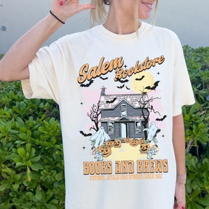 Op de afbeelding: Een cr&egrave;mekleurig T-shirt met een afbeelding van een spookhuis, geesten, vleermuizen en pompoenen. De tekst "Salem Bookstore" staat in een retro-lettertype boven het huis. Onder het huis staat de tekst "Books and Brews" met de slogan "Potions, Spells and Stories Since 1992".