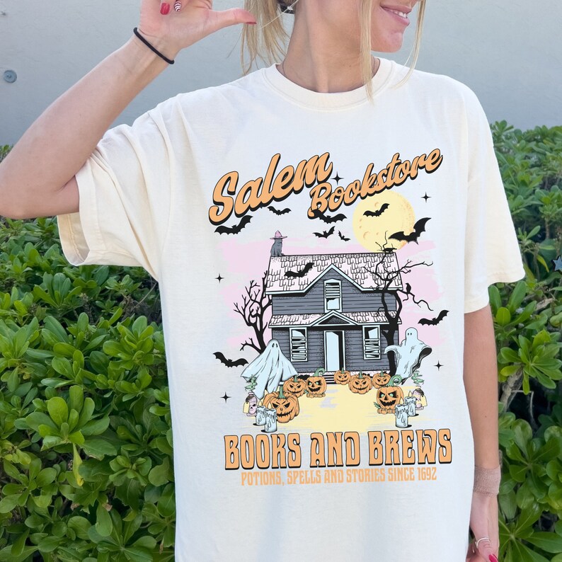 Op de afbeelding: Een cr&egrave;mekleurig T-shirt met een afbeelding van een spookhuis, geesten, vleermuizen en pompoenen. De tekst "Salem Bookstore" staat in een retro-lettertype boven het huis. Onder het huis staat de tekst "Books and Brews" met de slogan "Potions, Spells and Stories Since 1992".