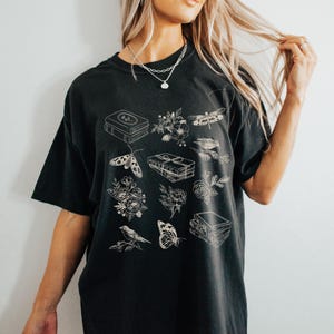 May include: Black t-shirt with a white graphic design featuring a stack of books, flowers, butterflies, dragonflies, and birds.