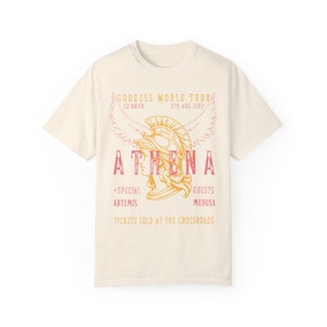 Puede incluir: Una camiseta de color crema con un gr&aacute;fico de estilo vintage de una mujer con un casco con alas. El texto dice "GODDESS WORLD TOUR", "ATHENA", "22:66NR", "5TN AUG.1982", "+SPECIAL ARTEMIS", "GUESTS MEDUSA", y "TICKETS SOLD AT THE CROSSROADS".