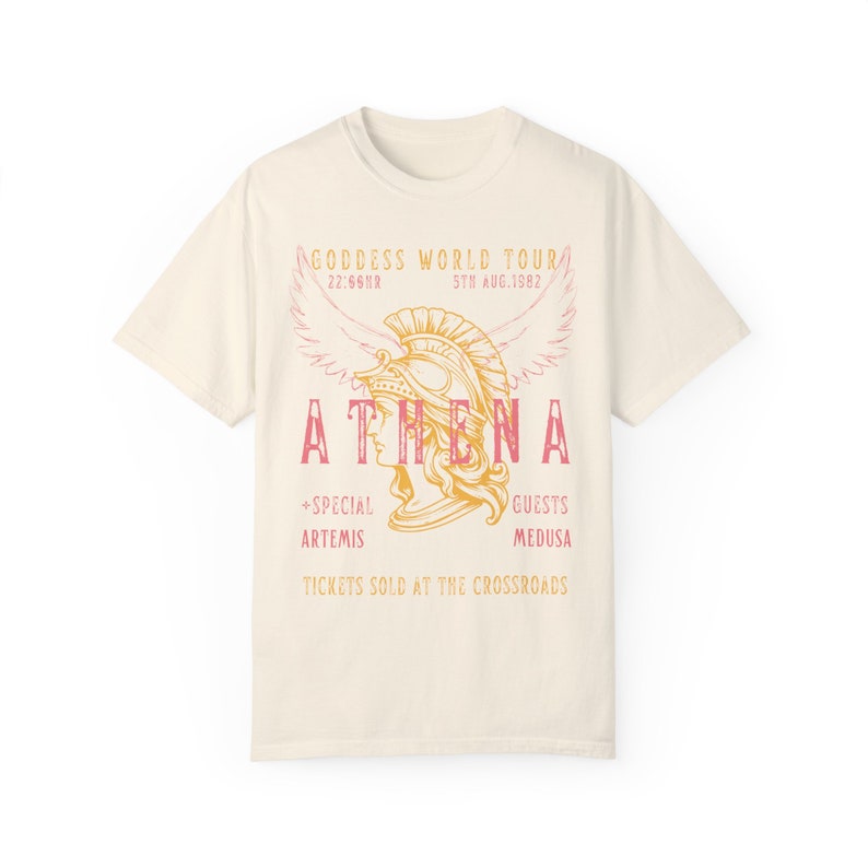 Puede incluir: Una camiseta de color crema con un gr&aacute;fico de estilo vintage de una mujer con un casco con alas. El texto dice "GODDESS WORLD TOUR", "ATHENA", "22:66NR", "5TN AUG.1982", "+SPECIAL ARTEMIS", "GUESTS MEDUSA", y "TICKETS SOLD AT THE CROSSROADS".