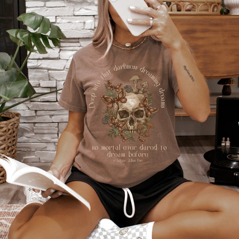 Pu&ograve; includere: Una maglietta marrone con un'immagine di un teschio circondato da farfalle e funghi. Il testo sulla maglietta recita "Deep into that darkness dreaming of dreams" e "no mortal ever dared to dream before - Edgar Allan Poe".