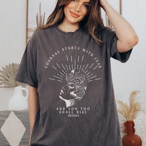 May include: A dark gray t-shirt with a white graphic print of a woman's face with snakes for hair. The text "Courage starts with fear and you too shall rise - Medusa" is printed below the graphic.
