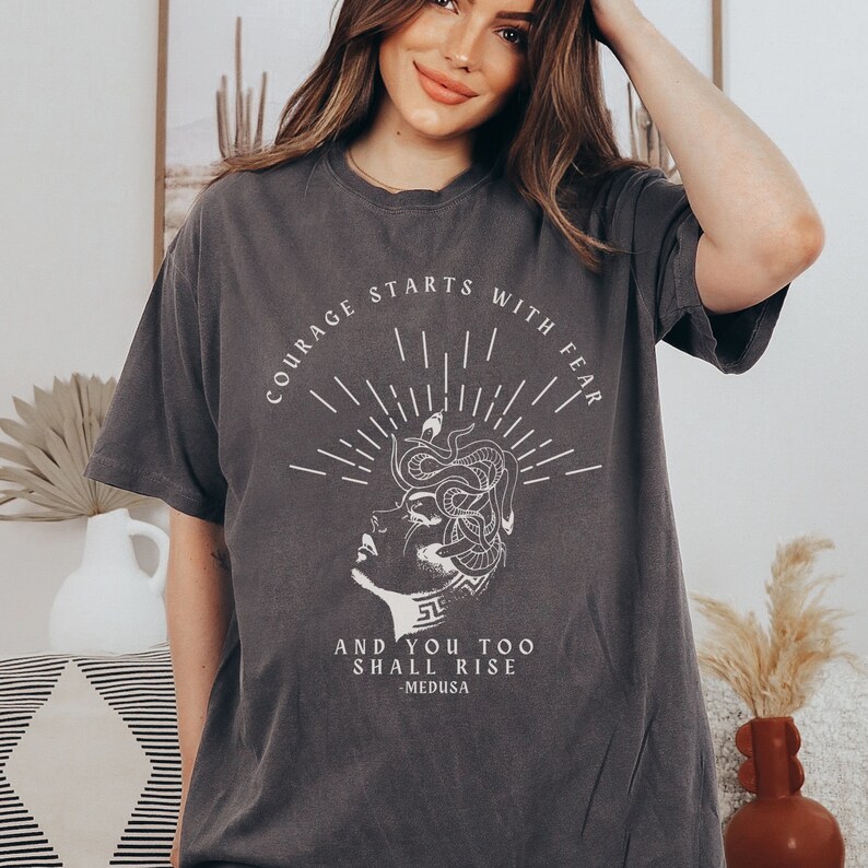 May include: A dark gray t-shirt with a white graphic print of a woman's face with snakes for hair. The text "Courage starts with fear and you too shall rise - Medusa" is printed below the graphic.
