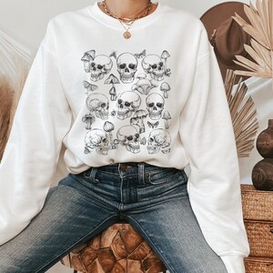 May include: White sweatshirt with a black and white print of skulls and mushrooms. The sweatshirt has long sleeves and a crew neck.