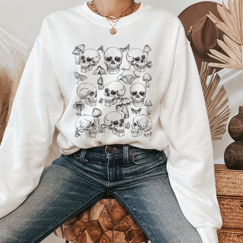 May include: White sweatshirt with a black and white print of skulls and mushrooms. The sweatshirt has long sleeves and a crew neck.