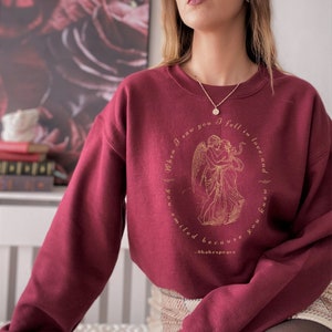 May include: Burgundy sweatshirt with a gold graphic of two angels embracing. The graphic is surrounded by text that reads "When I saw you I fell in love and  I smiled because you are no one" and "Shakespeare".
