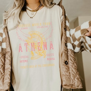 Puede incluir: Una camiseta blanca con un estampado gr&aacute;fico de una mujer con un casco con alas. El texto de la camiseta dice "Goddess World Tour 22:66HR 5TH AUG. 1982 ATHENA +SPECIAL ARTEMIS GUESTS MEDUSA TICKETS SOLD AT THE CROSSROADS".