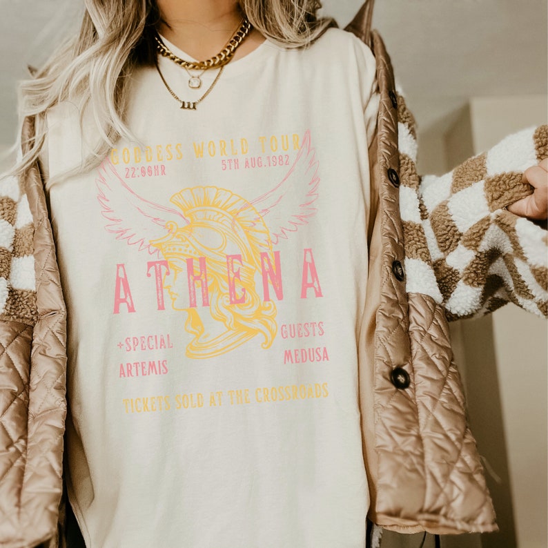 Puede incluir: Una camiseta blanca con un estampado gr&aacute;fico de una mujer con un casco con alas. El texto de la camiseta dice "Goddess World Tour 22:66HR 5TH AUG. 1982 ATHENA +SPECIAL ARTEMIS GUESTS MEDUSA TICKETS SOLD AT THE CROSSROADS".