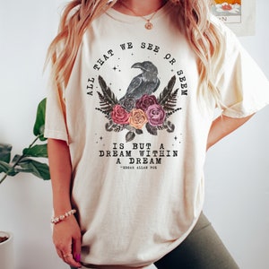 May include: A cream-colored t-shirt with a black raven and floral design. The text reads "All that we see or seem is but a dream within a dream - Edgar Allan Poe".