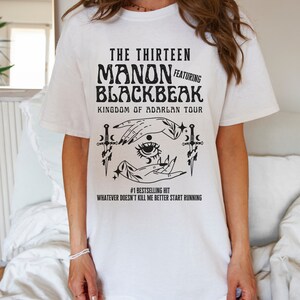 May include: White t-shirt with a black graphic design featuring the text "THE THIRTEEN MANON FEATURING BLACKBEAK KINGDOM OF ADARLAN TOUR" and a stylized illustration of two hands with an eye in the centre, surrounded by stars and daggers. The text "#1 BESTSELLING HIT WHATEVER DOESN'T KILL ME BETTER START RUNNING" is below the illustration.