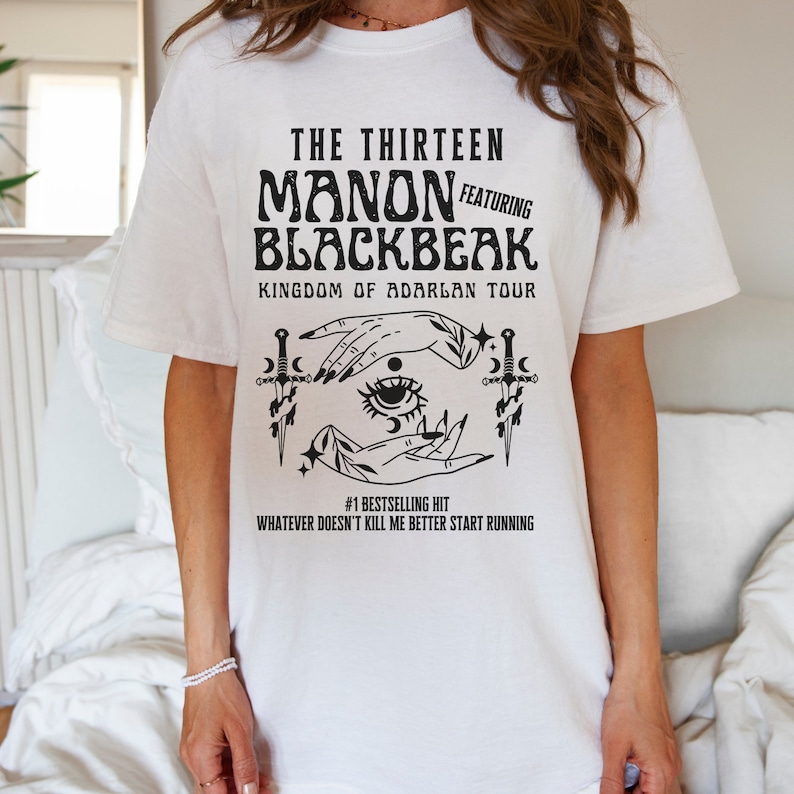 May include: White t-shirt with a black graphic design featuring the text "THE THIRTEEN MANON FEATURING BLACKBEAK KINGDOM OF ADARLAN TOUR" and a stylized illustration of two hands with an eye in the centre, surrounded by stars and daggers. The text "#1 BESTSELLING HIT WHATEVER DOESN'T KILL ME BETTER START RUNNING" is below the illustration.