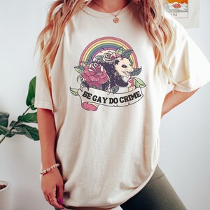 May include: Cream-colored t-shirt featuring a colorful graphic. The design includes a rainbow, roses, and an opossum. A banner below the image reads "BE GAY DO CRIME". The shirt has short sleeves and a relaxed fit.