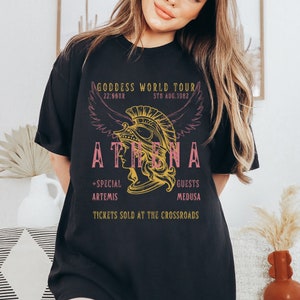 Puede incluir: Camiseta negra con un dise&ntilde;o gr&aacute;fico rosa y amarillo que representa a una mujer con un casco con alas. El texto dice "Goddess World Tour", "22:00HR", "5TH AUG.1982", "ATHENA", "+SPECIAL ARTEMIS", "GUESTS MEDUSA" y "TICKETS SOLD AT THE CROSSROADS".