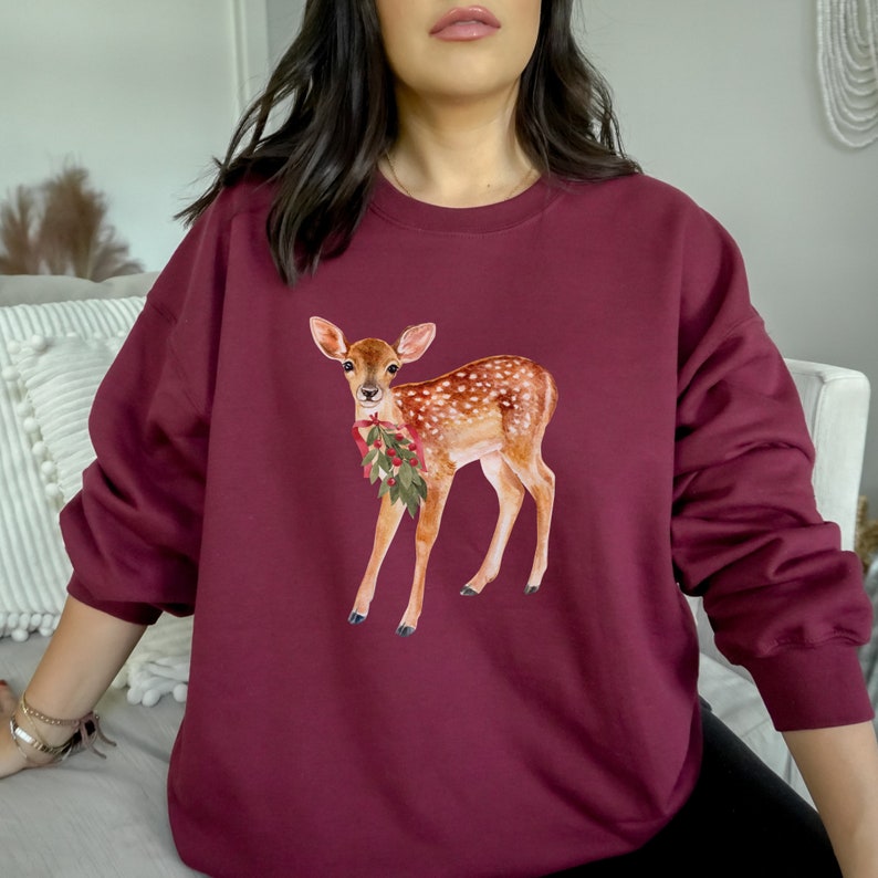 May include: A maroon sweatshirt with a watercolor illustration of a fawn wearing a red and green holly sprig. The fawn is standing on all four legs and looking to the left.