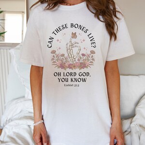 Puede incluir: Camiseta blanca con un gr&aacute;fico negro de una mano esquel&eacute;tica sosteniendo una mariposa. El texto dice "Can these bones live? Oh Lord God, you know Ezekiel 37:3".