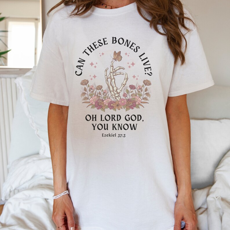 Puede incluir: Camiseta blanca con un gr&aacute;fico negro de una mano esquel&eacute;tica sosteniendo una mariposa. El texto dice "Can these bones live? Oh Lord God, you know Ezekiel 37:3".