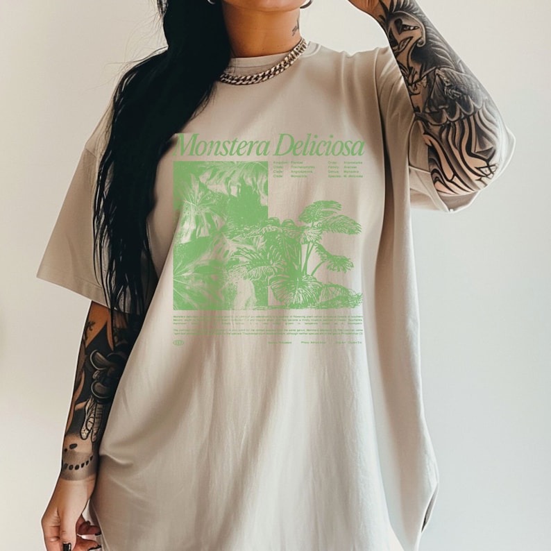 May include: A beige oversized t-shirt with a green graphic print of a Monstera Deliciosa plant. The graphic includes the text "Monstera Deliciosa" and a description of the plant.