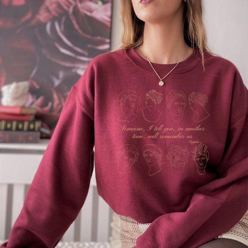 Pode incluir: Camisola de cor bord&ocirc; com um contorno dourado de v&aacute;rios rostos femininos e o texto "Algu&eacute;m, eu te digo, em outro tempo, se lembrar&aacute; de n&oacute;s - Safo".